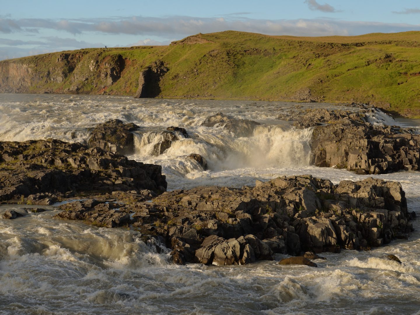 Rocky waterfall cascades powerfully over dark stones, surrounded by green hills under a blue sky, conveying a sense of rugged natural beauty.