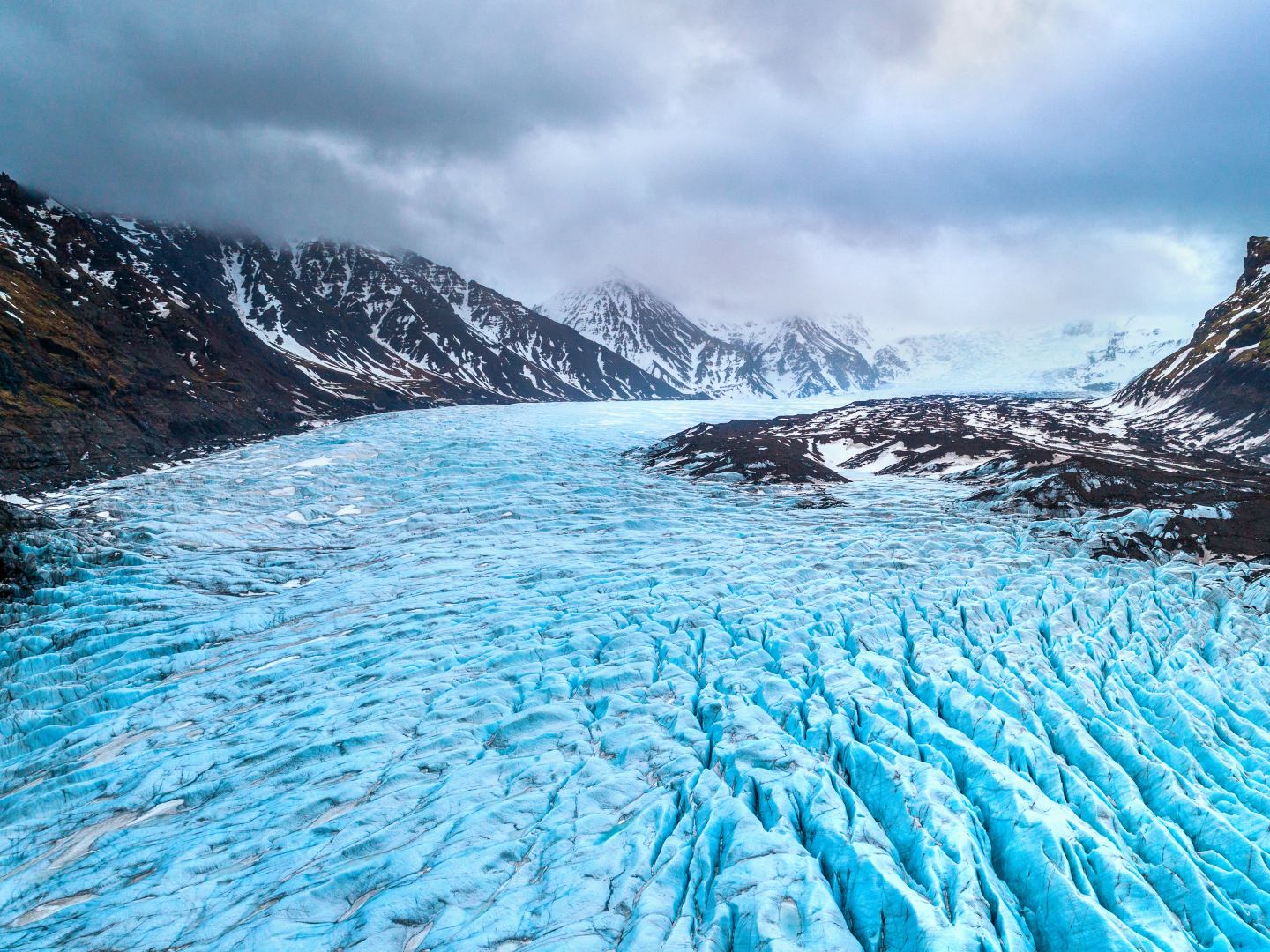 Expansive blue glacier nestled between rugged, dark mountains under a cloudy sky. The scene conveys a sense of cold, remote wilderness.