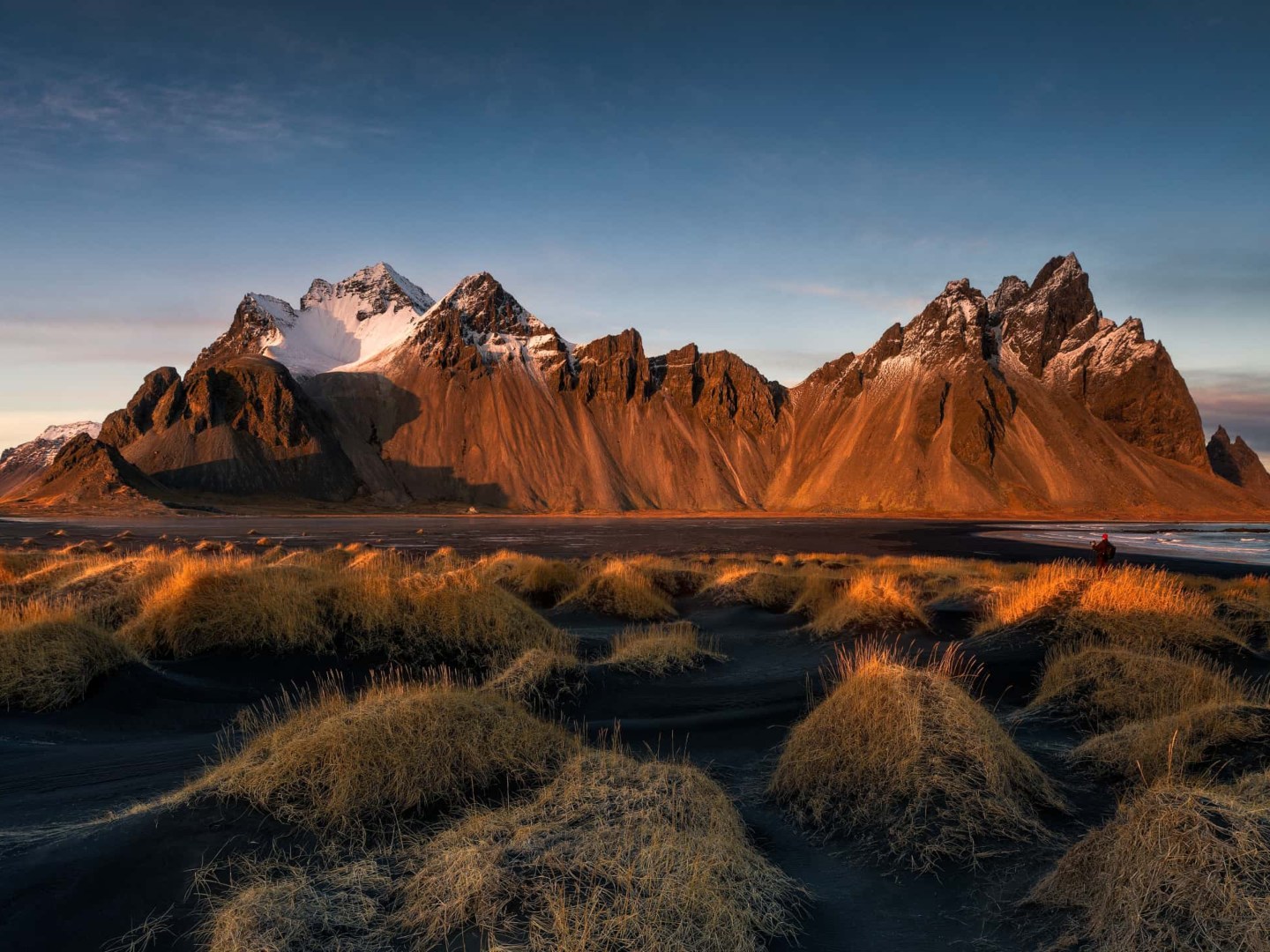 Golden grass tufts cover dark dunes in the foreground, with jagged, snow-capped mountains glowing in warm sunset light under a clear sky.