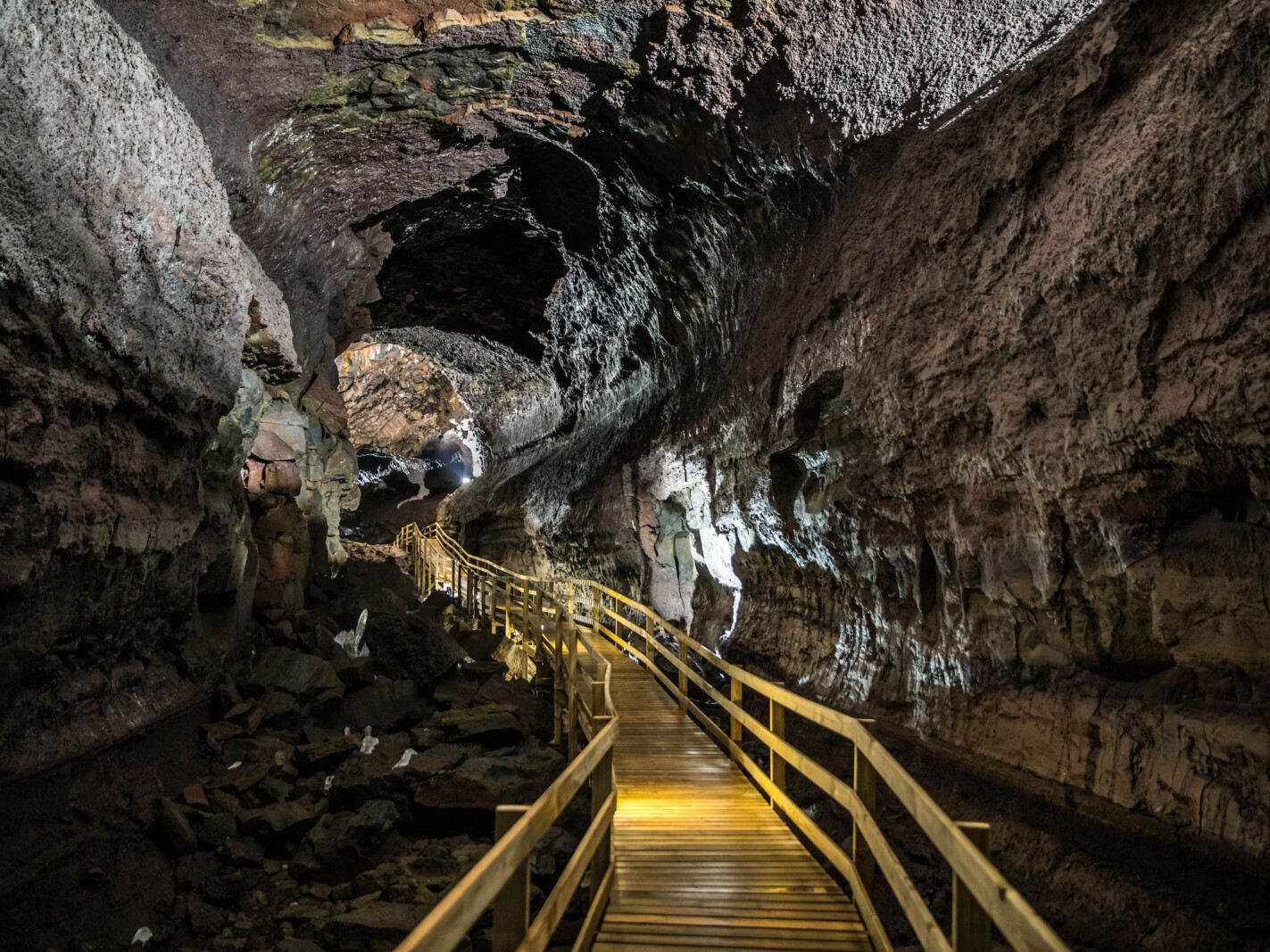 A well-lit wooden walkway winds through a dark, rocky cave. The textured walls and ceiling are illuminated, creating a mysterious and adventurous atmosphere.