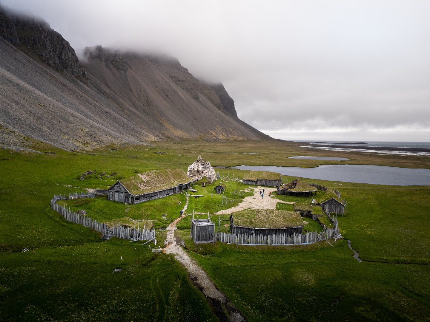 Viking village replica on lush green grass under a cloudy sky, with wooden structures and fences. A mountain looms in the background, adding a serene feel.
