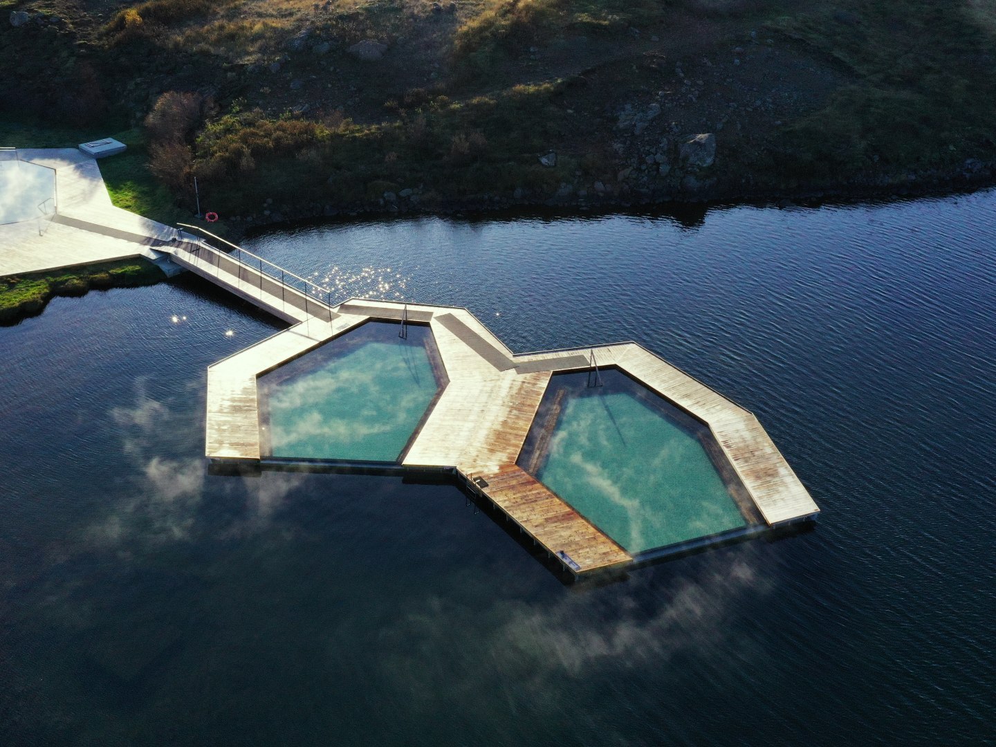 Aerial view of two connected hexagonal pools with wooden decks, floating on a calm lake surrounded by rocky terrain. Mist rises, adding a serene ambiance.