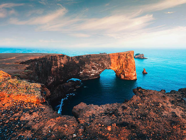 Rocky coastal arch over blue ocean in Iceland, with dramatic cliffs under a cloudy sky. Warm sunlight casts a serene glow on the rugged landscape.