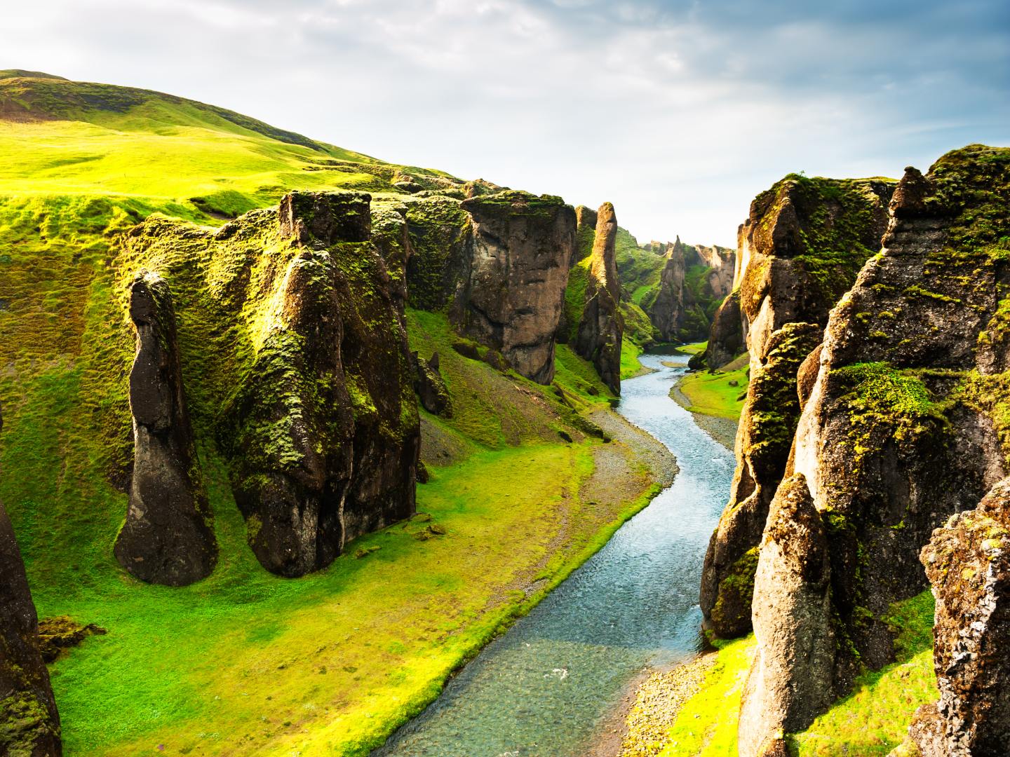 A river winds through a lush, green canyon with towering moss-covered cliffs under a partly cloudy sky, evoking a sense of tranquility and awe.
