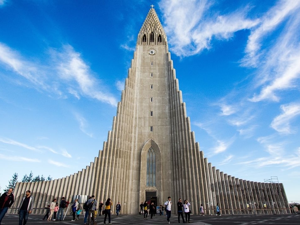 Towering concrete church with a triangular facade, clock, and people outside. Clear blue sky with scattered clouds, conveying a majestic and serene atmosphere.