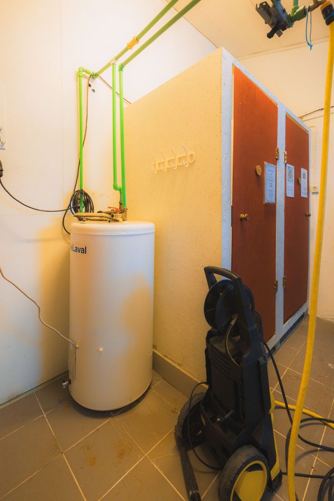 Room with a large white tank, green pipes overhead, and a black-yellow pressure washer on the tiled floor. Adjacent, a wooden cubicle with hooks.