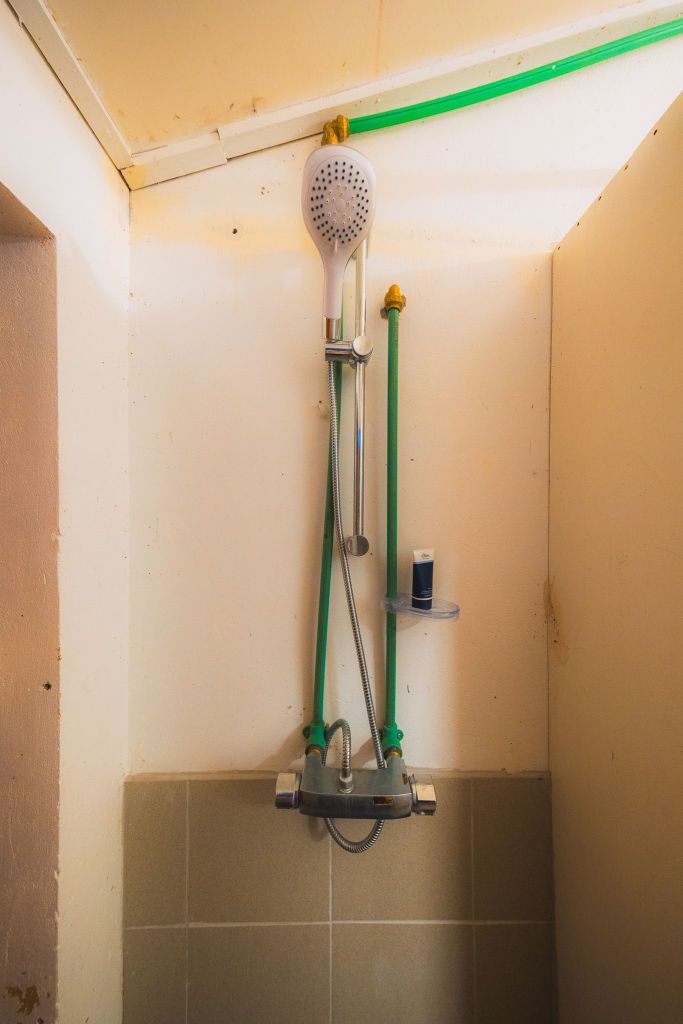 Shower with a white head, green pipes, and a gray hose against a beige tiled wall. A small shelf holds a blue bottle, creating a basic and functional look.