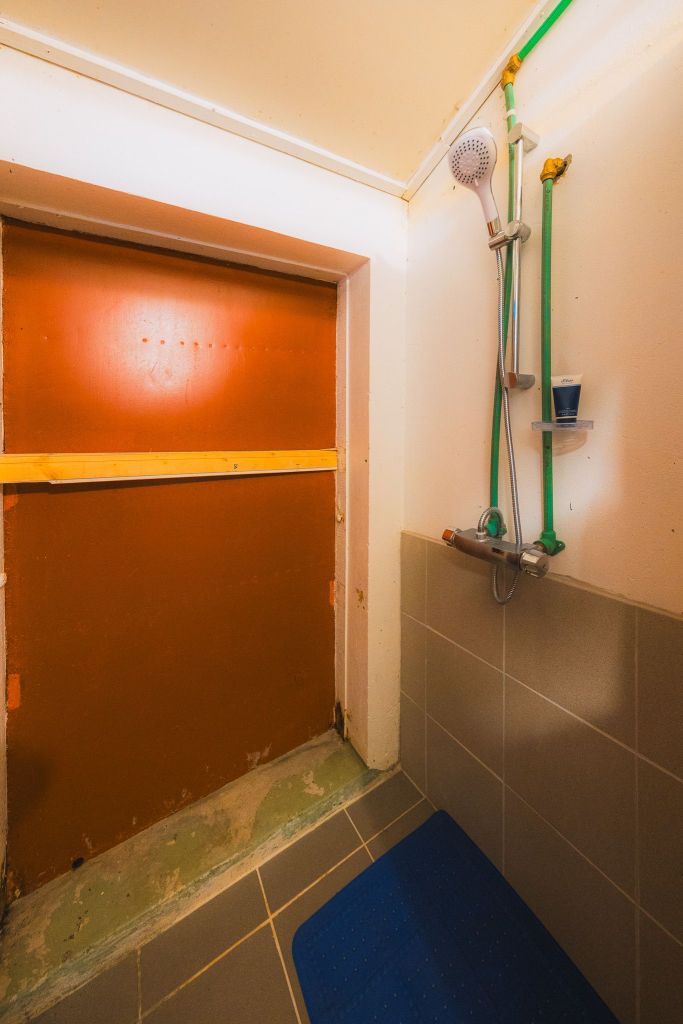 A small shower area with beige tiles, a gray showerhead, and green piping. A blue mat covers the floor, and a brown boarded-up window is on the left.