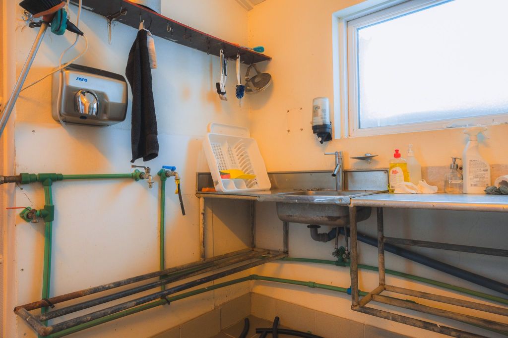 A utility room featuring a stainless steel sink, dish rack, cleaning supplies, and a towel. Pipes run along the walls, under a bright window.