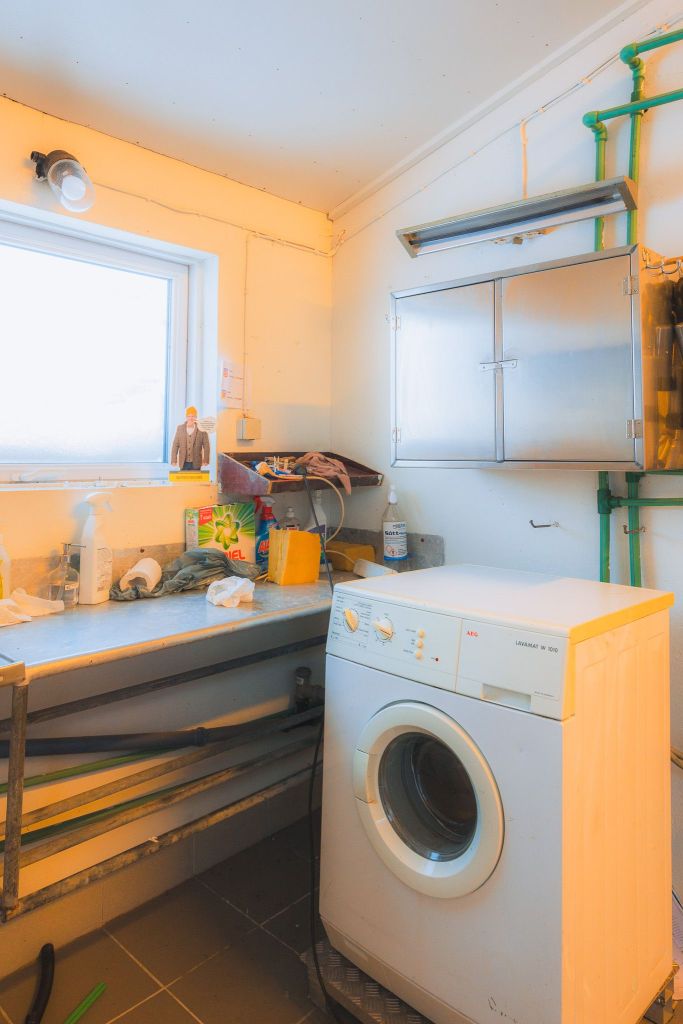 A small laundry room with a white washing machine beside a cluttered counter. Cleaning supplies, pipes, and a small window are visible.