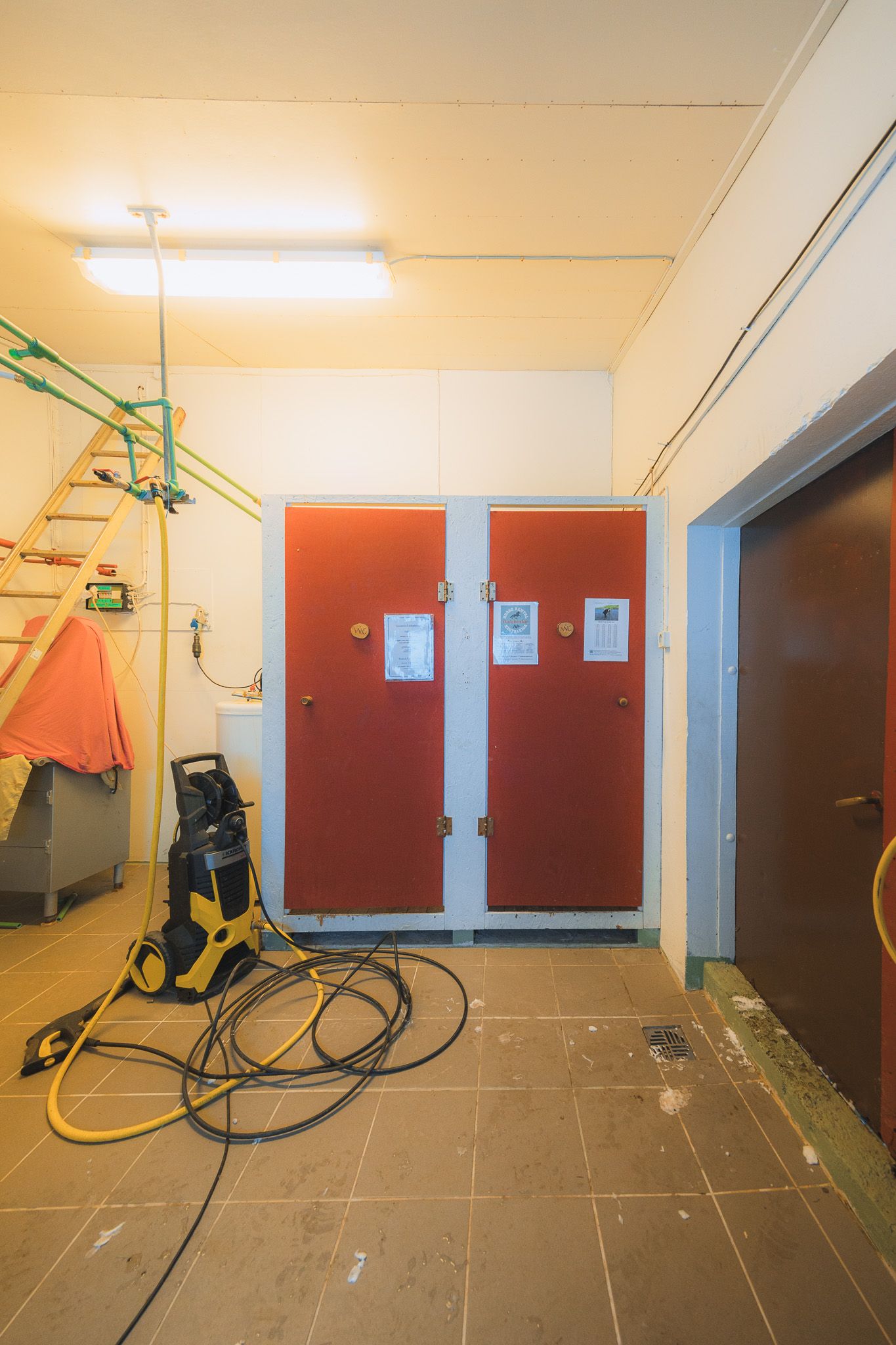 A dimly lit utility room with red double doors, a pressure washer with tangled hoses on the tiled floor, and a ladder on the left wall.