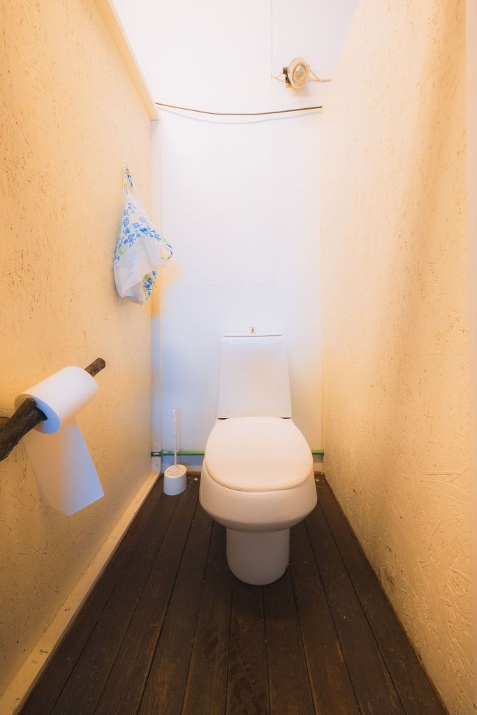 Narrow bathroom with a white toilet against an off-white wall. A roll of toilet paper is on a rustic holder, and cleaning supplies are on the wooden floor.