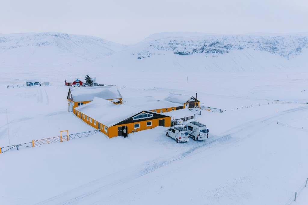 Aerial view of a large, yellow building with snowy roofs, set in an expansive, white, wintry landscape with distant mountains. Two white camper vans are parked nearby.