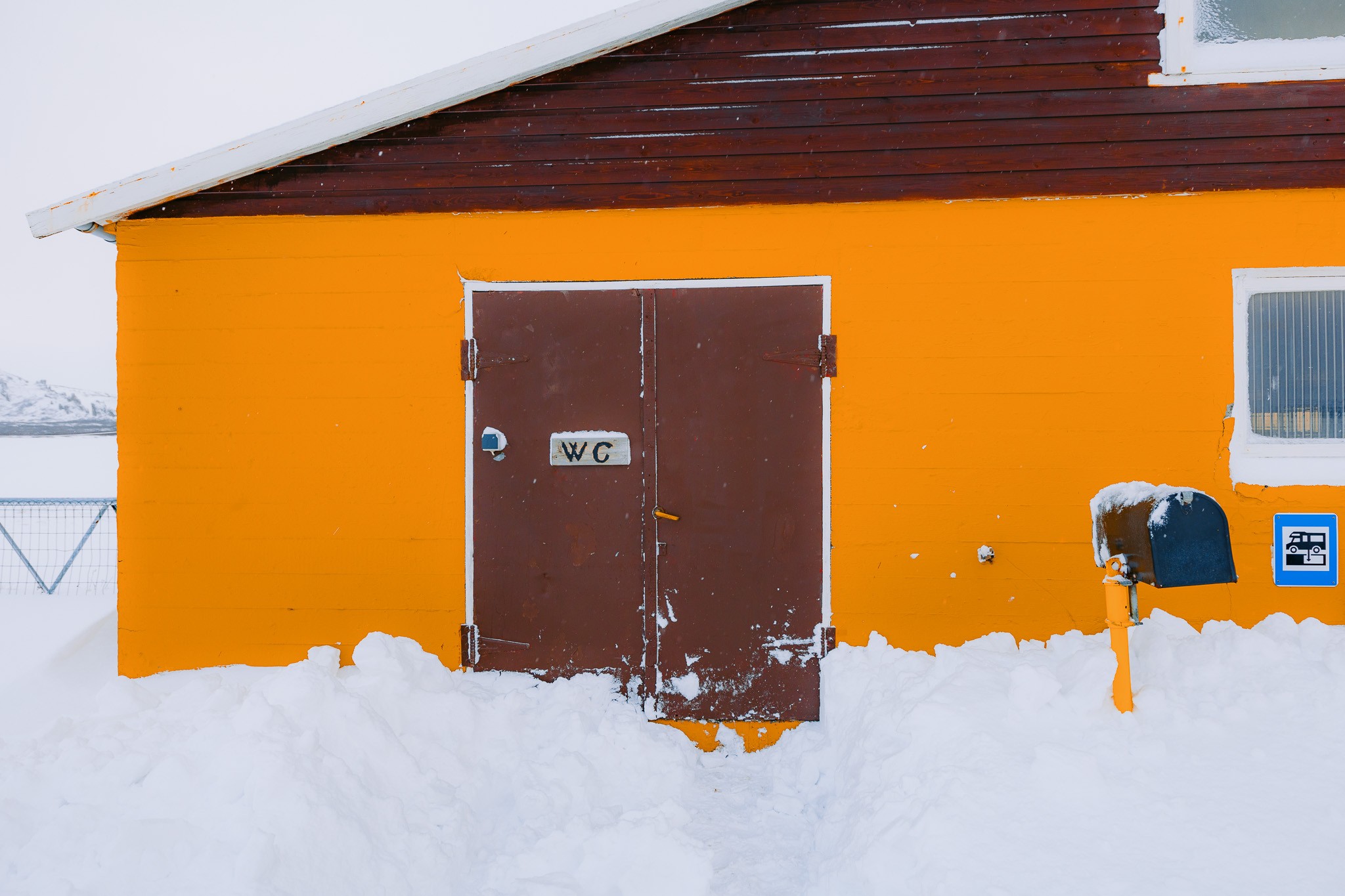 A vivid orange building with a snow-covered entrance showing a brown door marked "WC."