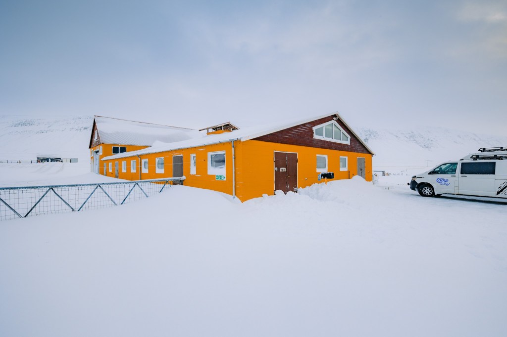 A bright yellow building stands against a snowy landscape with a cloudy sky. A white campervan is parked nearby.