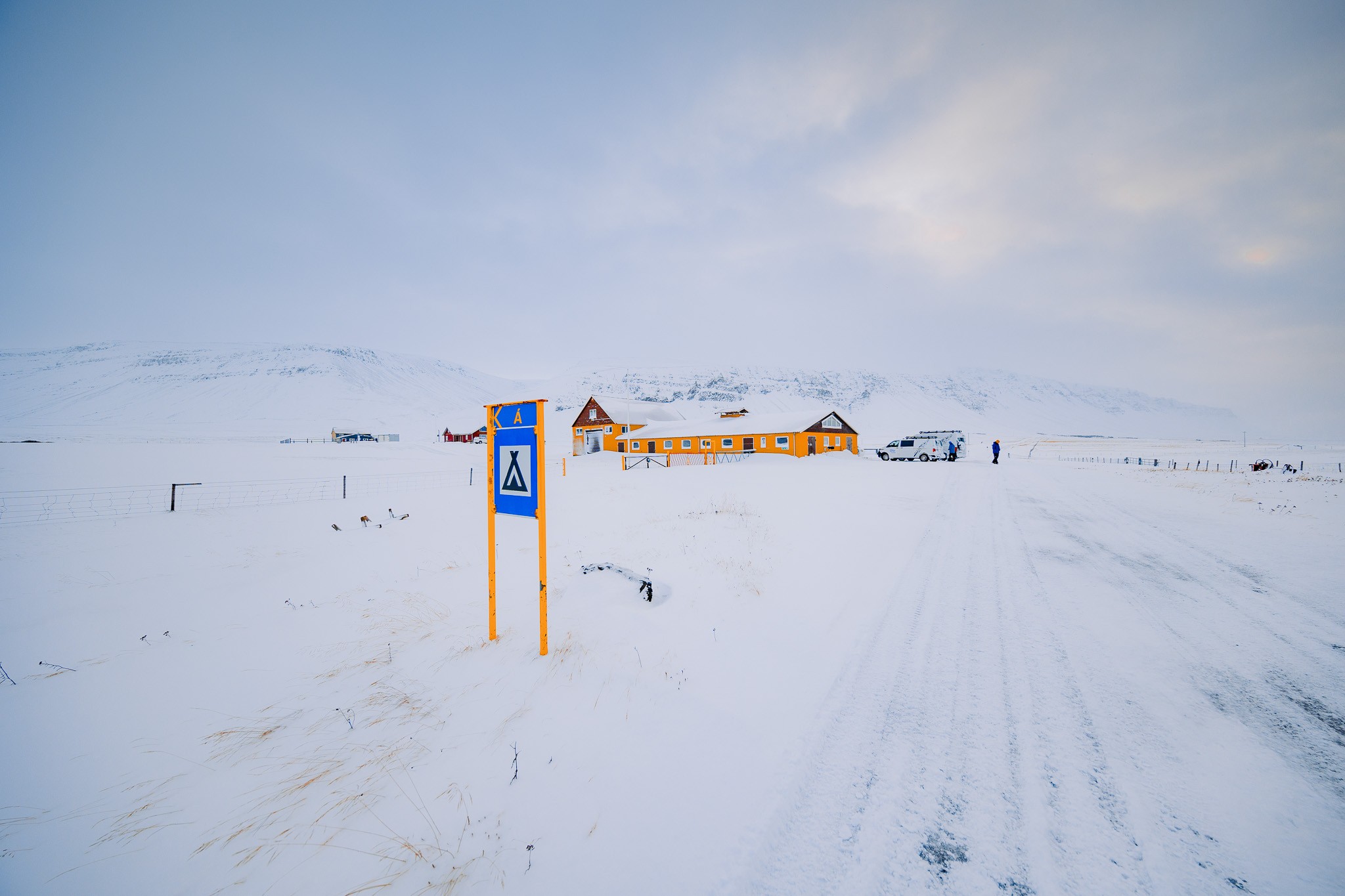 Snow-covered landscape with a bright orange building in the distance. A blue camping sign is in the foreground.