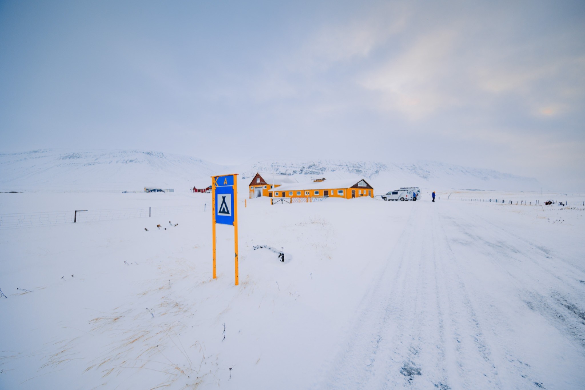 Snow-covered landscape with a bright orange building in the distance. A blue camping sign is in the foreground.