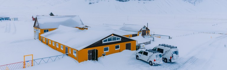 A snowy landscape with bright orange buildings, partially covered in snow, and two white vehicles nearby.