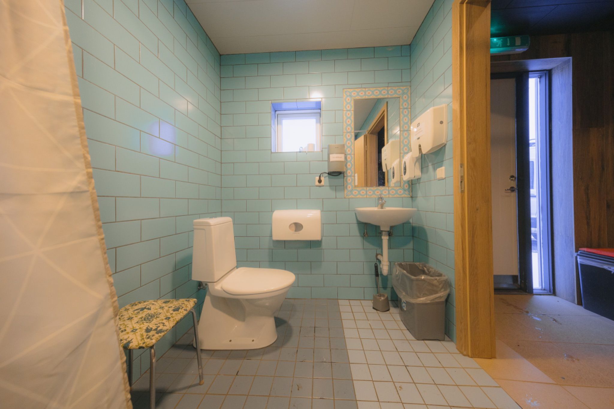 A clean bathroom features light blue tiles, a white toilet, and a sink with a mirror. A curtain to the left.