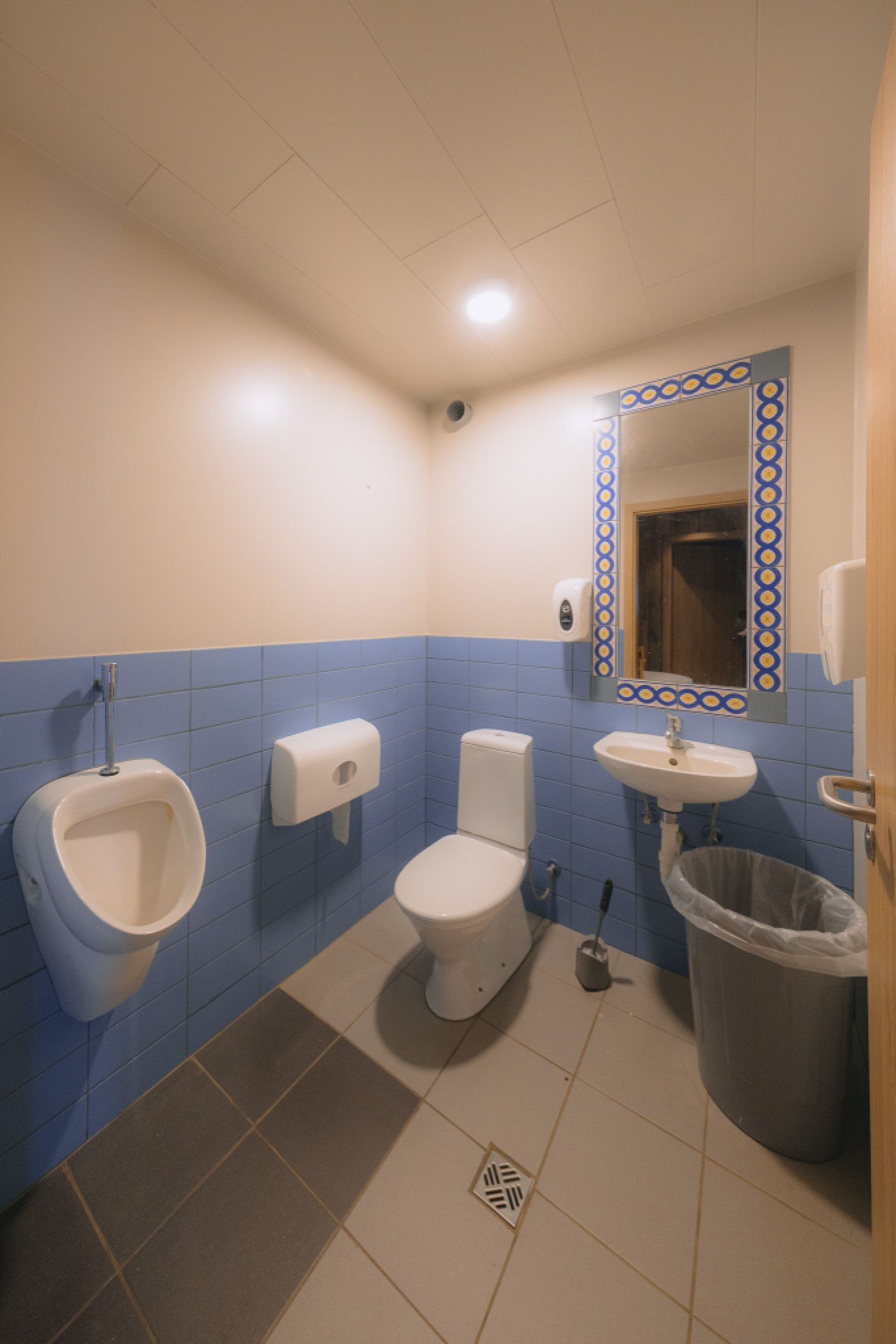 Small restroom with blue-tiled walls, urinal, toilet, and sink. A patterned mirror hangs above the sink, next to a large trash can.