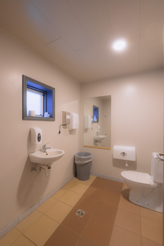 Clean restroom with beige walls, a small sink with a soap dispenser, and a toilet. A mirror and trash can are nearby, with a small window above.