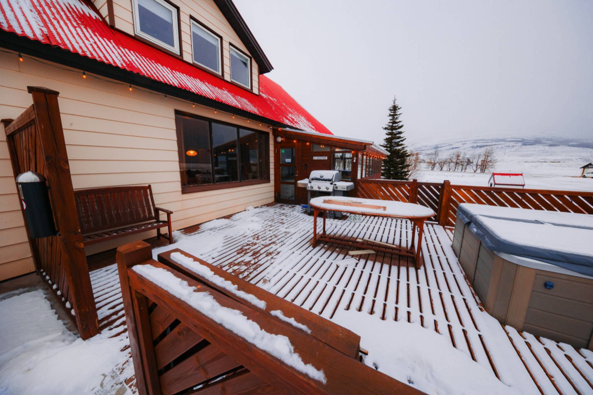 Snow-covered deck with wooden furniture and a hot tub outside a beige house with a bright red roof.