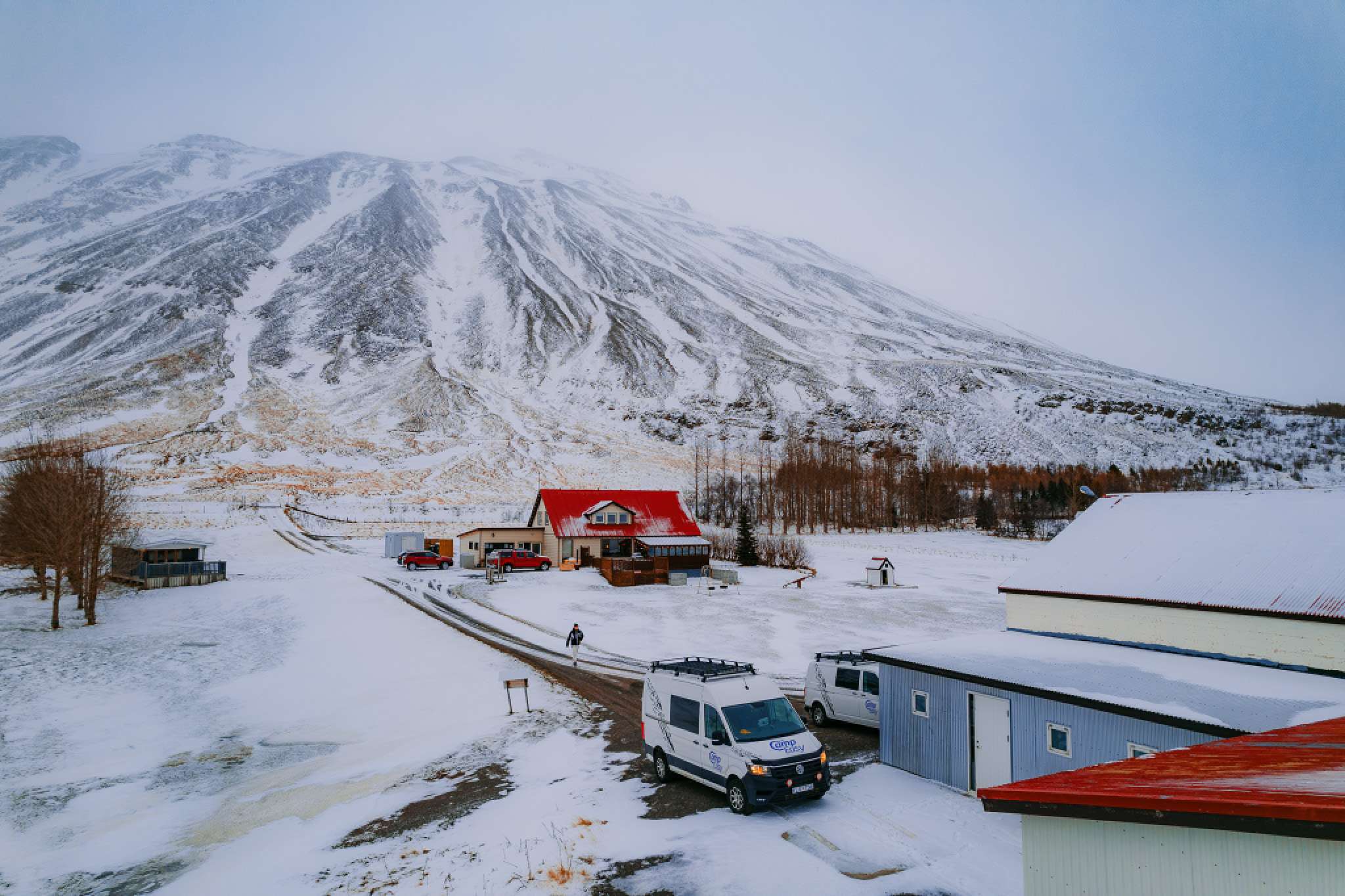 A snowy landscape with a large mountain in the background, small buildings with red roofs, and a campervans parked on a snow-covered road.