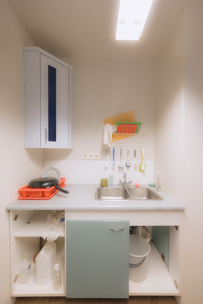 Small kitchen area with a double sink, filled dish rack, and various utensils hanging above. Open cupboard below shows cleaning items.