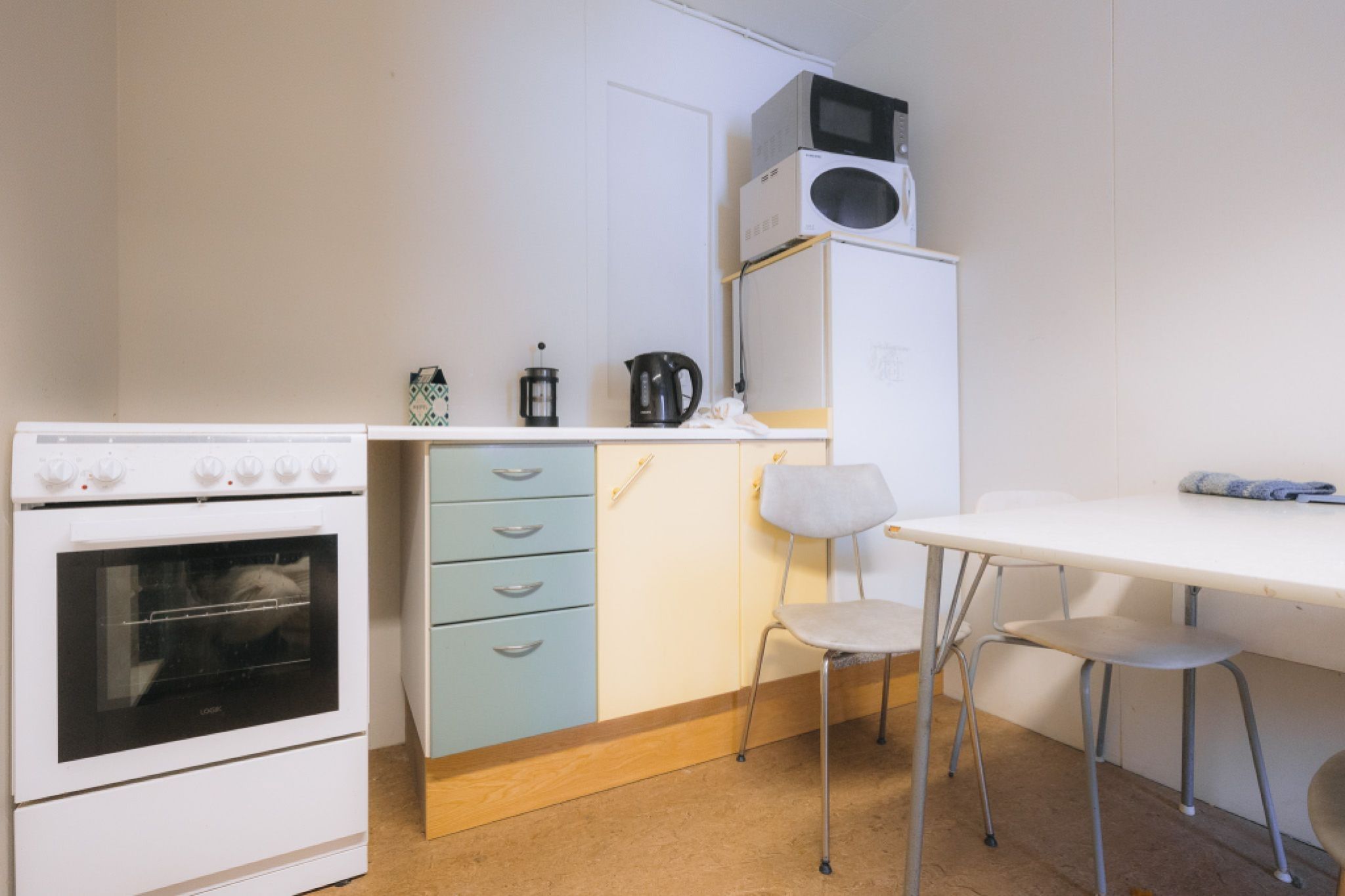 Compact kitchen with a stove, kettle, French press, and stacked fridge with microwave. Table and chairs next to it.