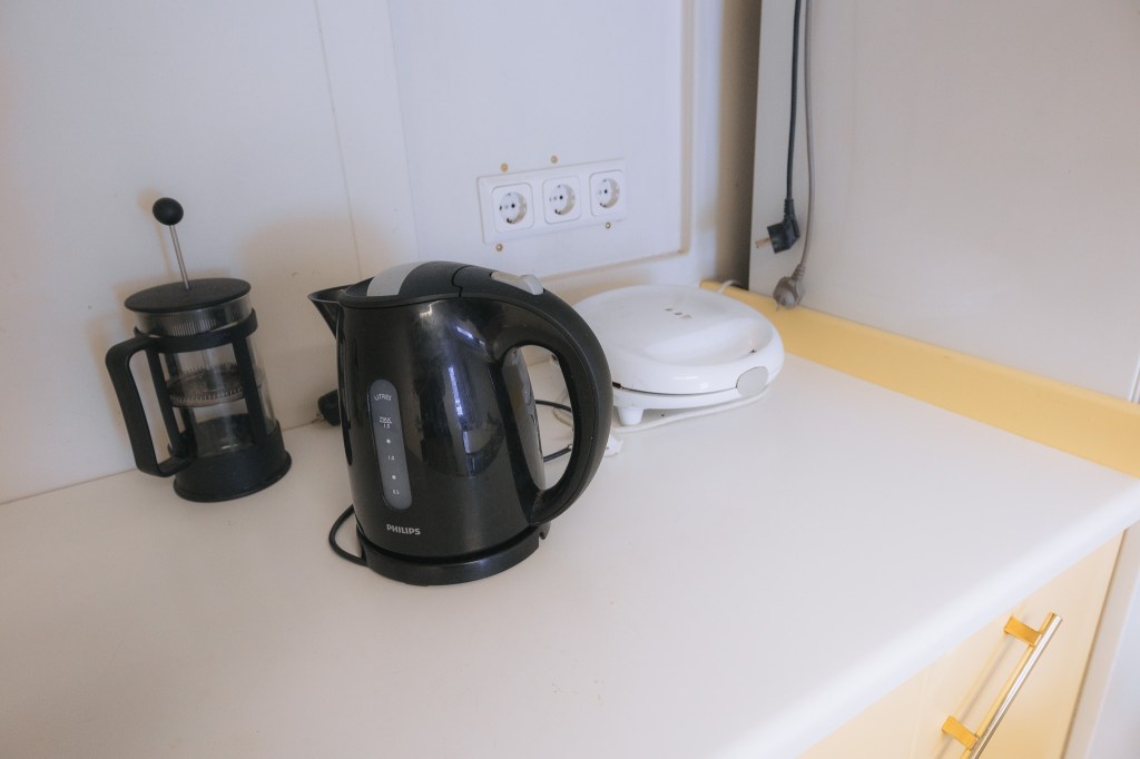 Electric kettle, French press, and sandwich maker on a kitchen counter. A power outlet is on the wal.
