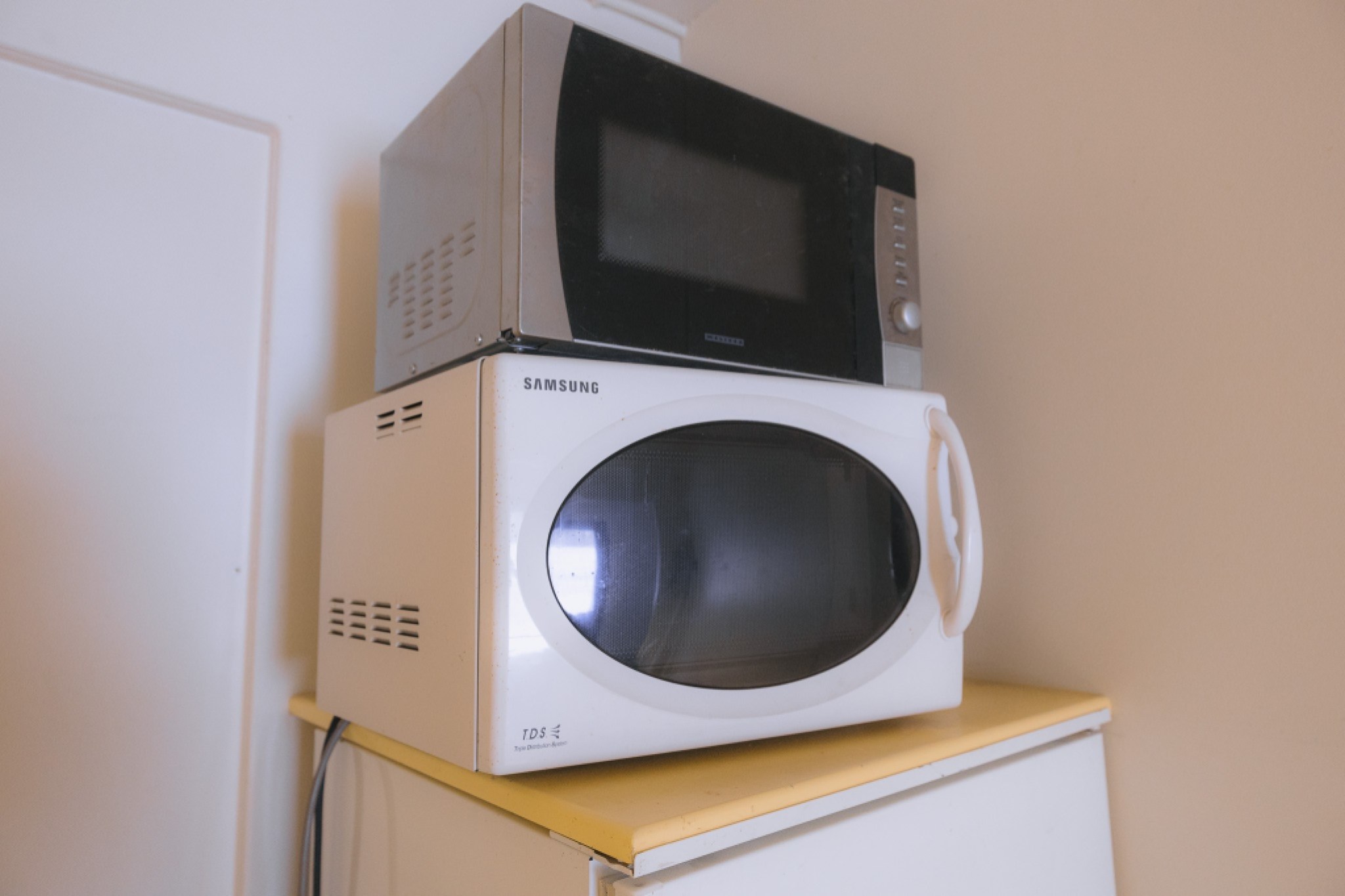 Two stacked microwaves on a refrigerator: a black and stainless upper microwave, and a white Samsung with a curved handle below, in a plain kitchen corner.