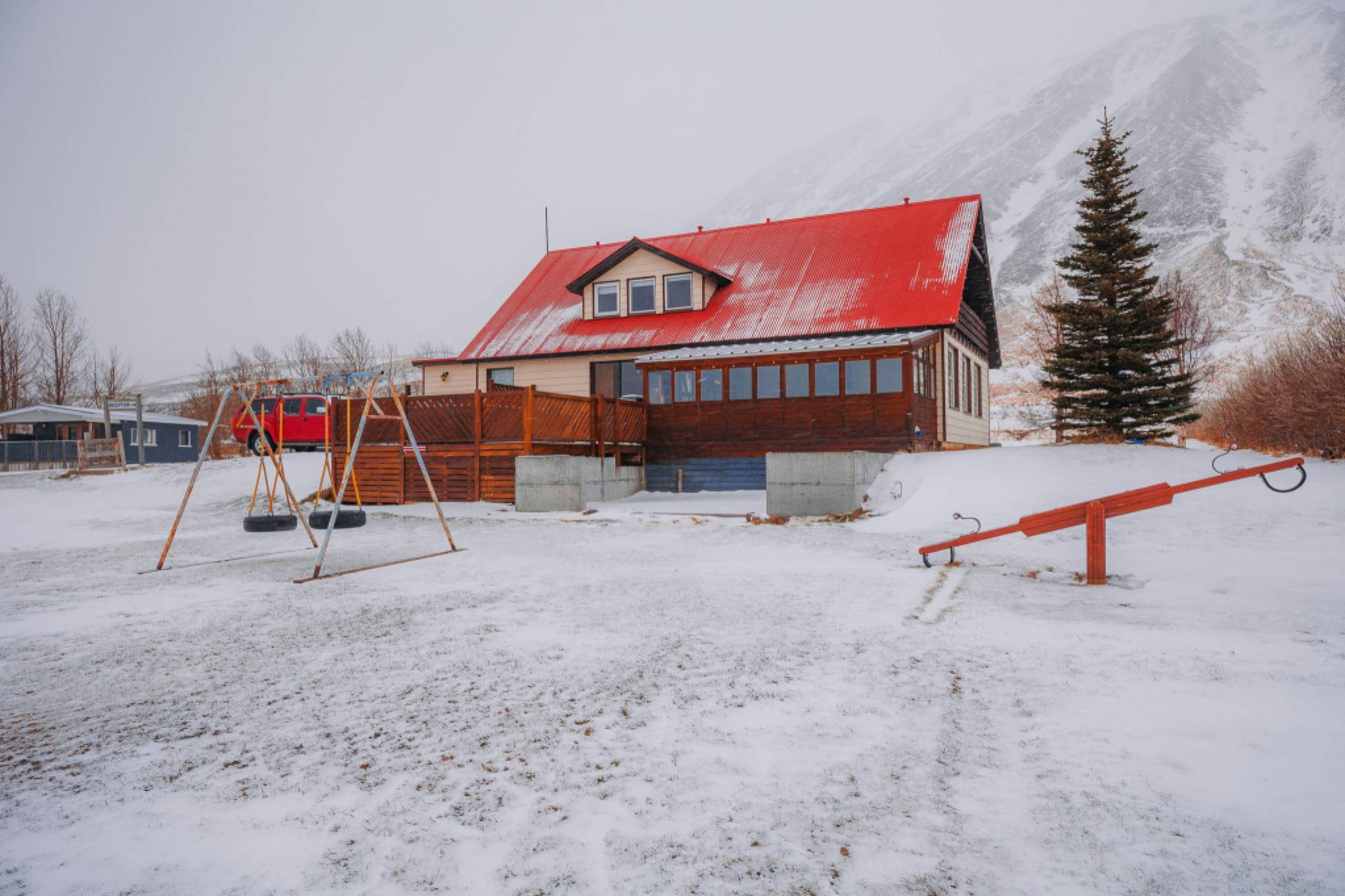 A wooden house with a red roof stands in a snowy landscape, with a swing set and a see-saw in front.