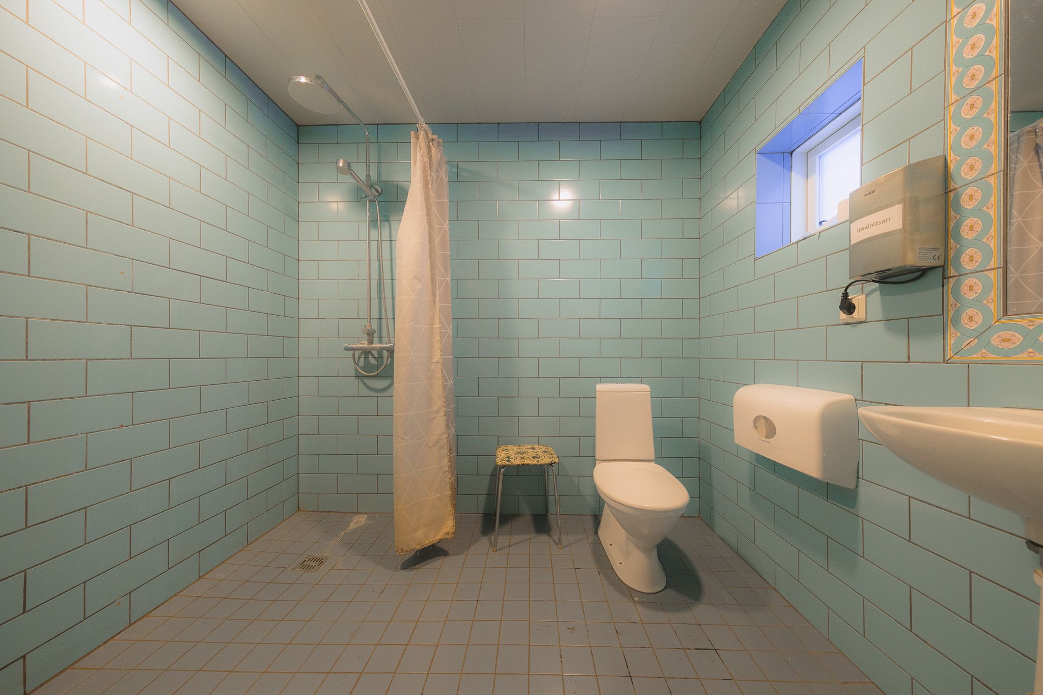 Spacious bathroom with light blue tiles, featuring a shower with a curtain, a toilet, a small stool, a sink, and a mirror.