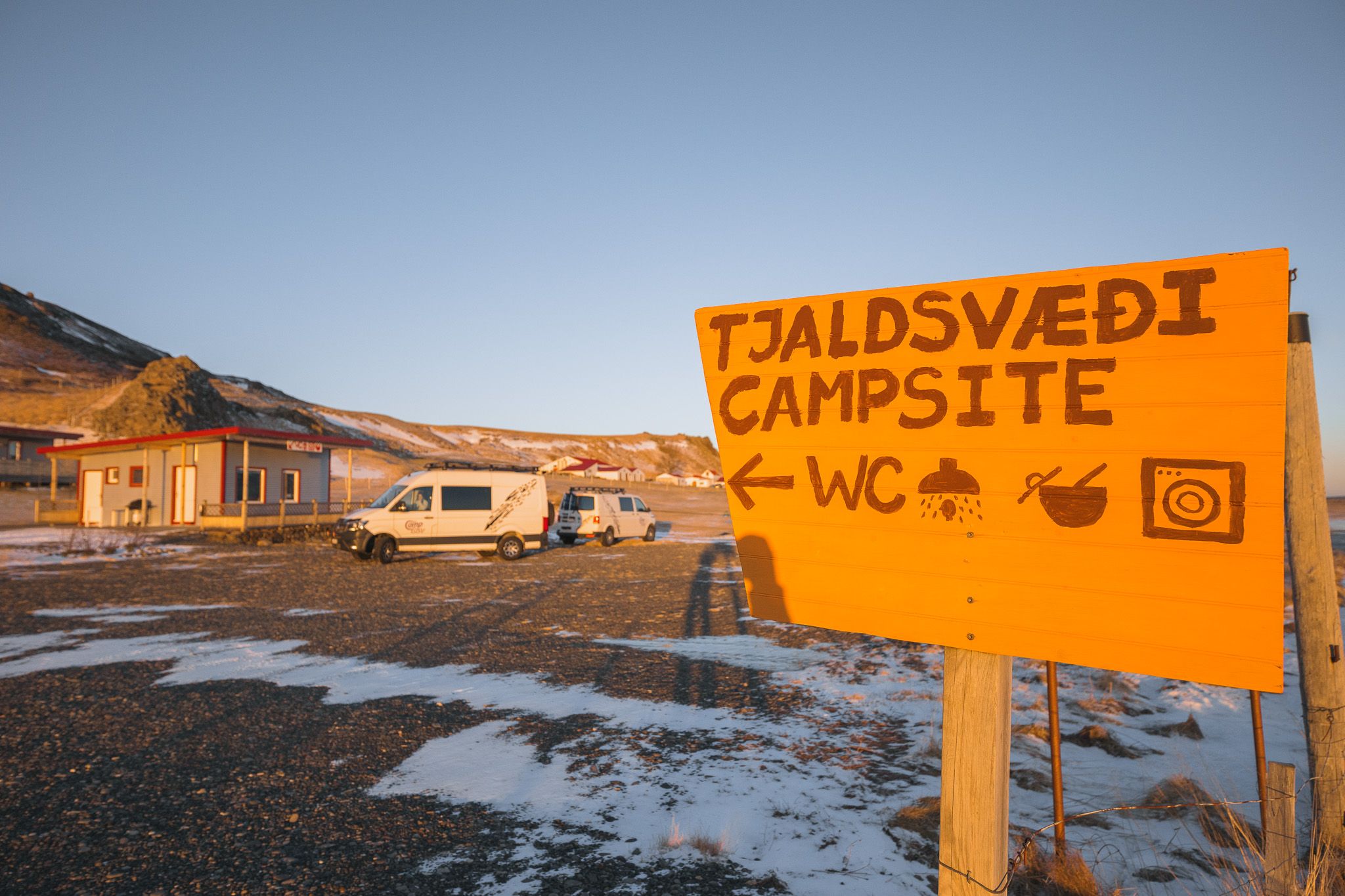 Sunlit campsite with a bright yellow sign in the foreground reading "Tjaldsvæði Campsite." Nearby are parked vans and a small building. Snow patches cover the ground.