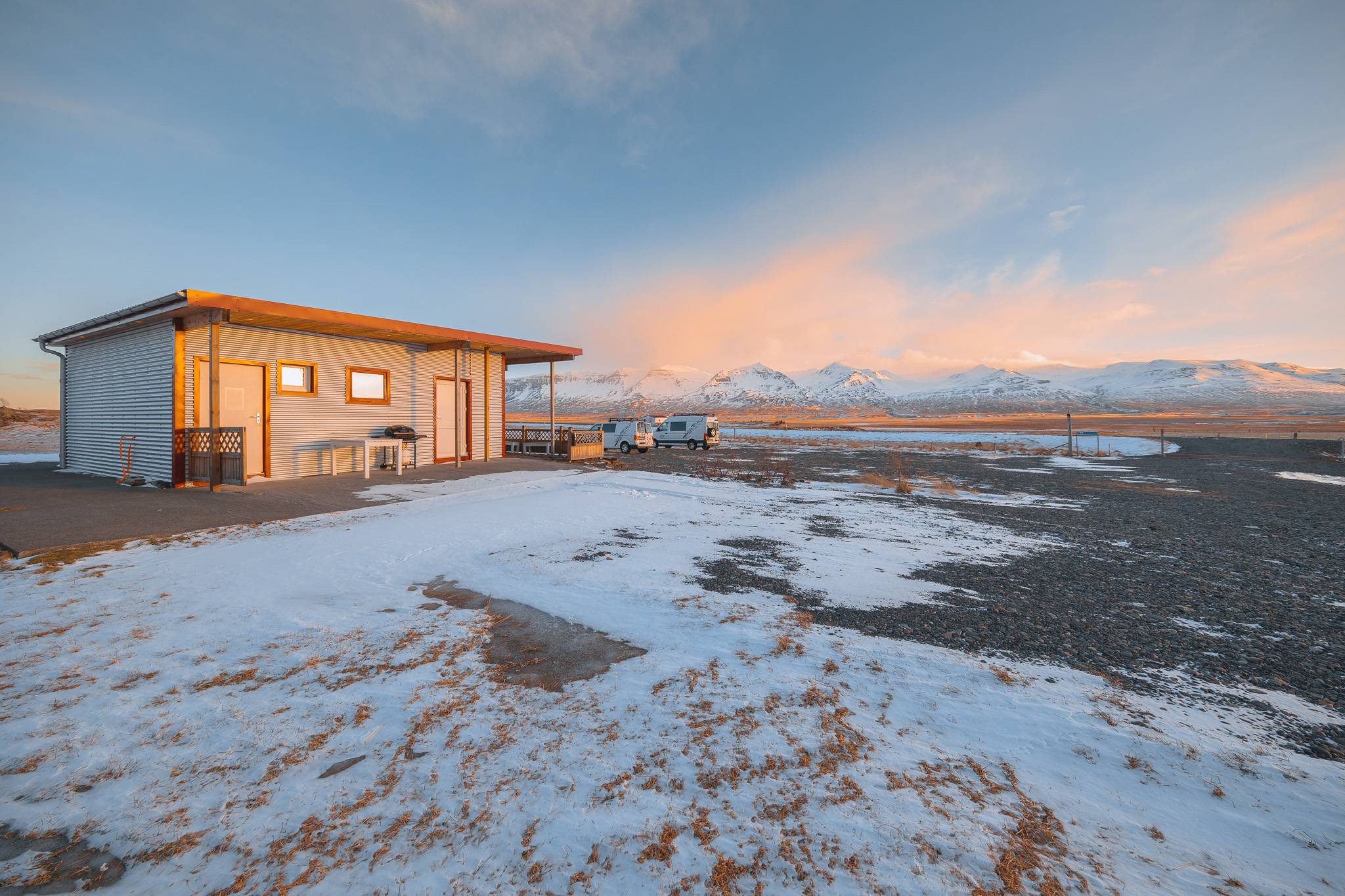 A small house stands on a snowy landscape with mountains in the background under a pink sunset sky.
