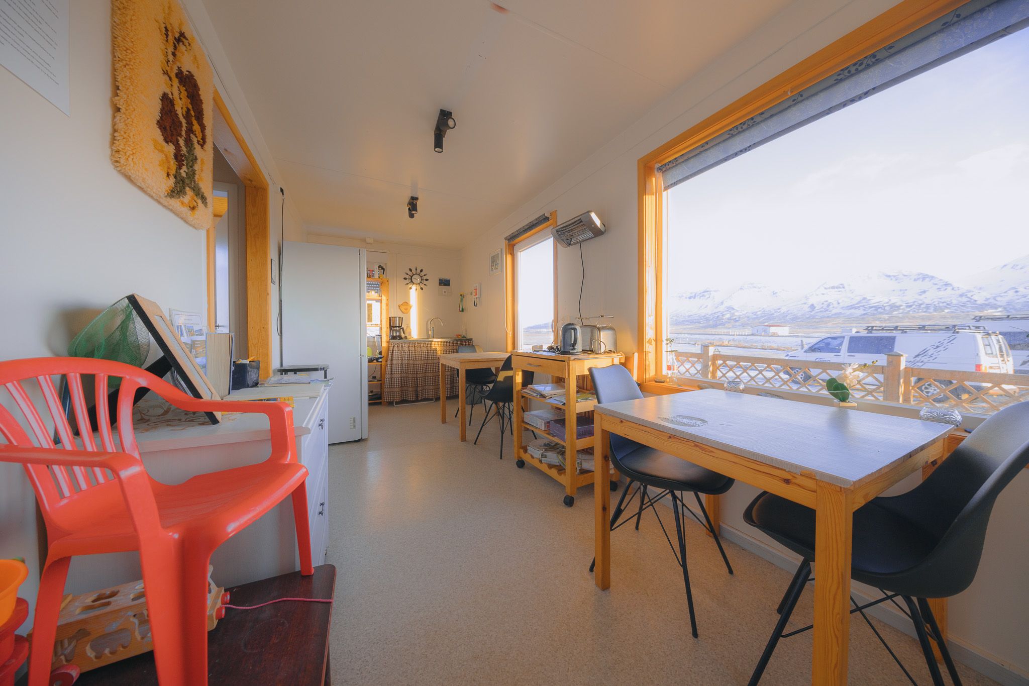 Cozy, sunlit dining area with large windows framing snowy mountains. Features wooden tables, black chairs, red chair, and wicker decor.