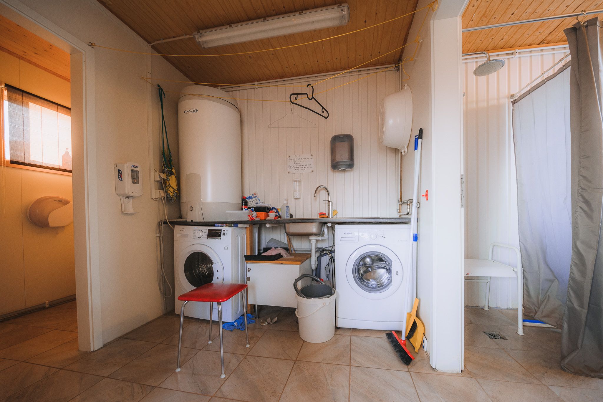 Laundry room with a cozy, functional setup. Two washing machines sit under a counter; a red stool and cleaning supplies add color.