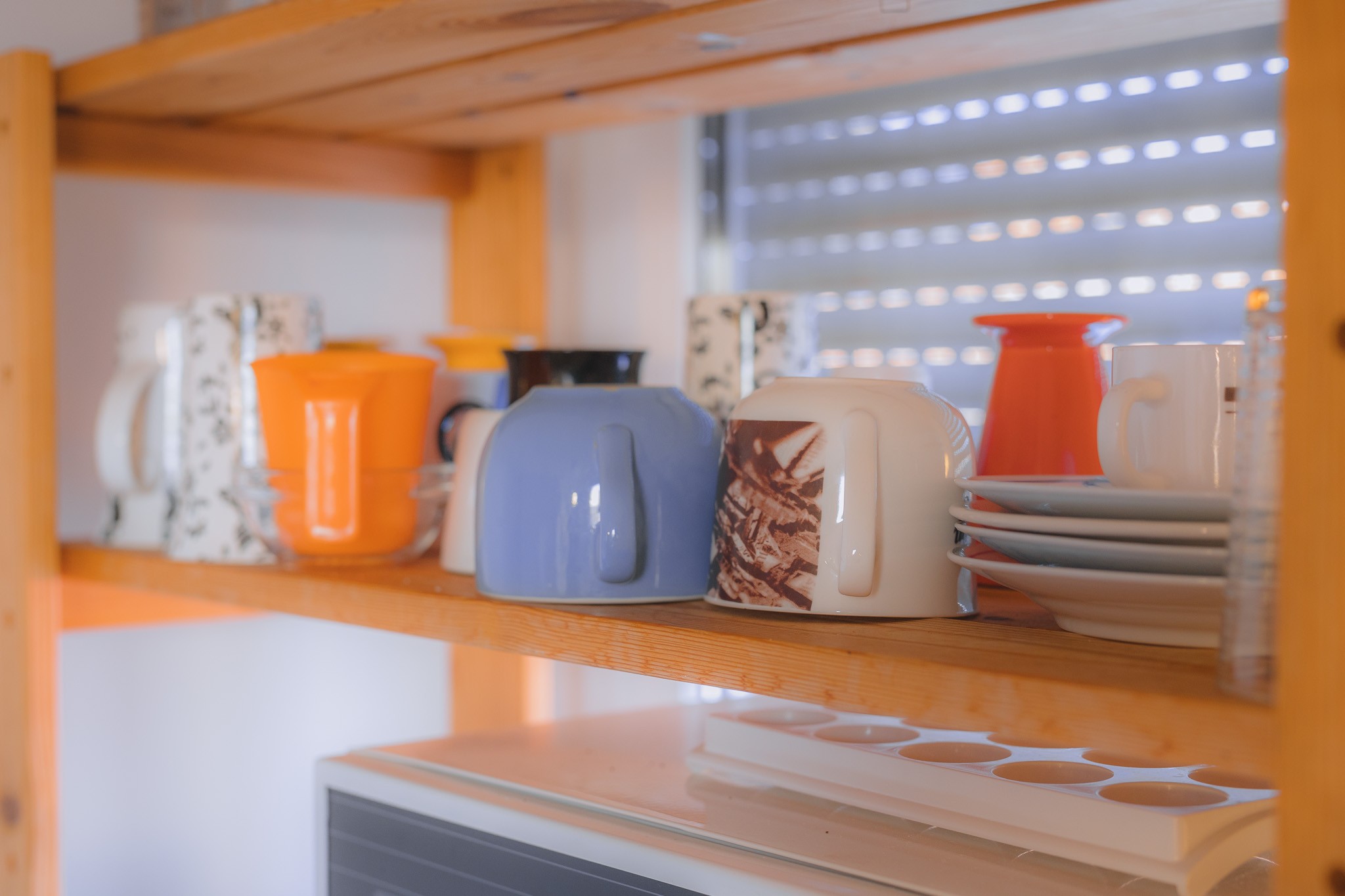 Wooden kitchen shelf holding colorful mugs and stacked plates, including orange, blue, and patterned designs, with soft, warm lighting.