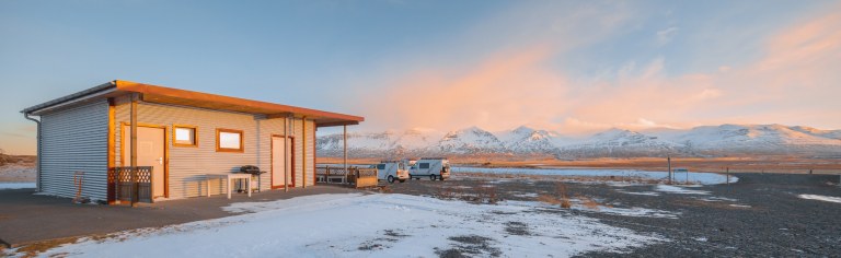 A small cabin stands in a snowy landscape under a vast sky with pink clouds. Mountains line the horizon, and a van is parked nearby, conveying solitude.