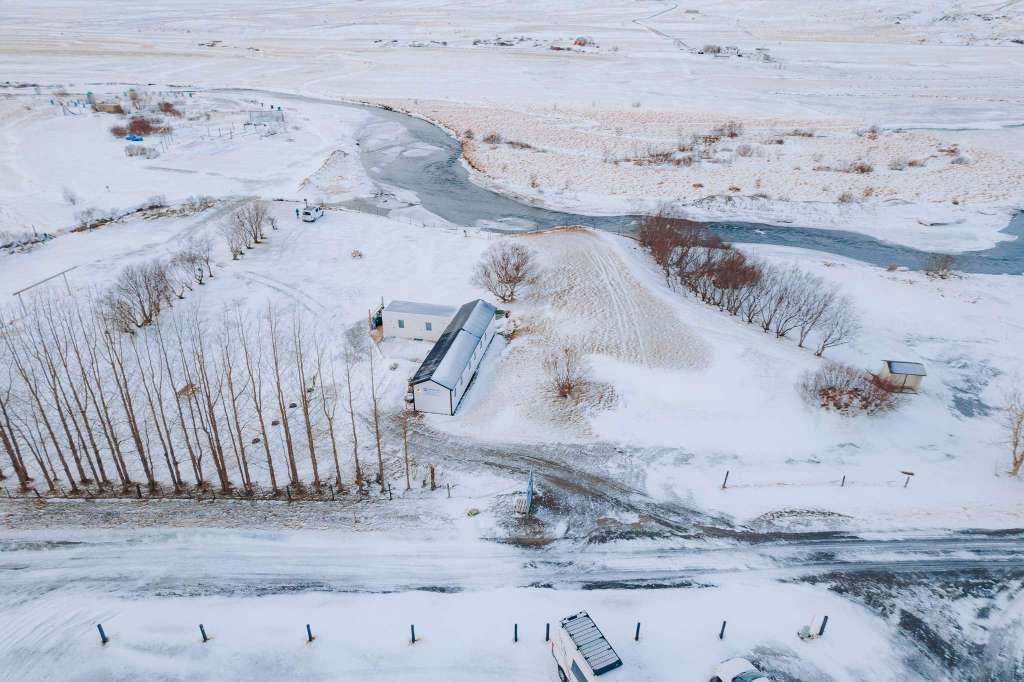 Aerial view of a snowy rural landscape with two buildings near a winding river. Rows of leafless trees line the road.