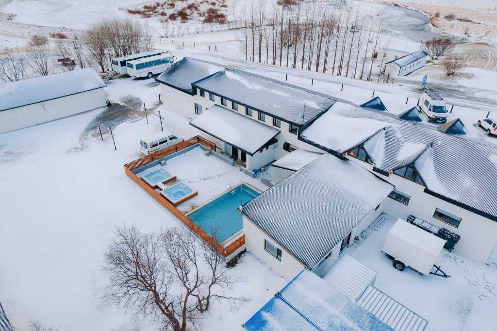 Snow-covered hotel with outdoor pool and hot tubs, surrounded by a snowy landscape. Nearby, vehicles are parked, and bare trees line the road.