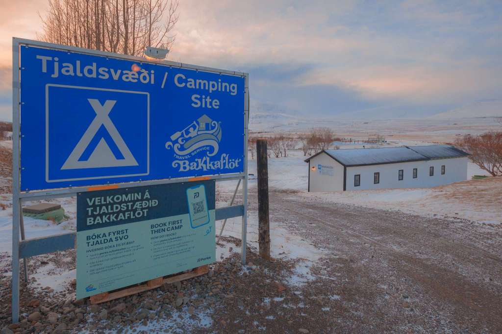 Snowy campsite scene with a large blue “Camping Site” sign in the foreground. A white building is set against a tranquil winter landscape.