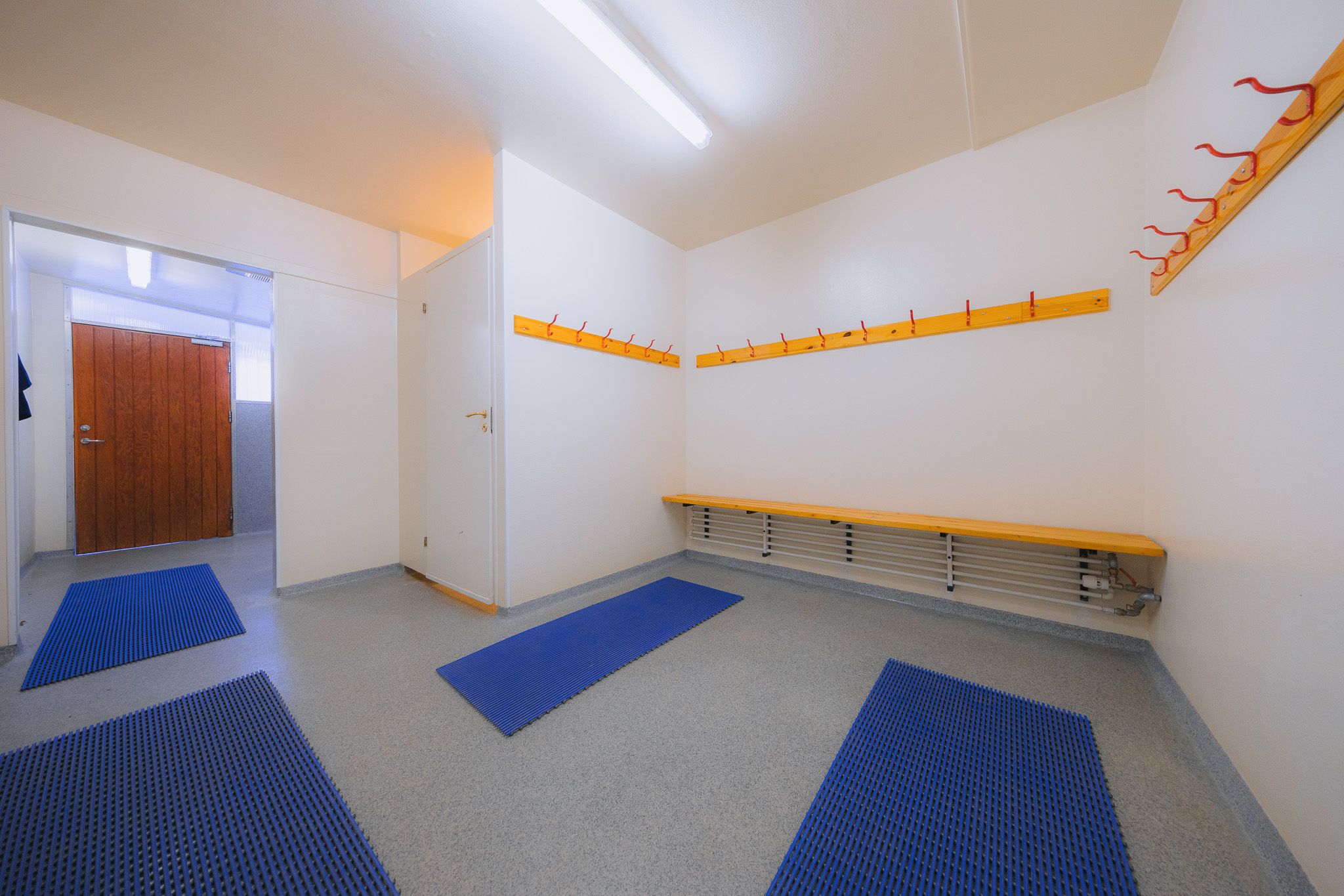 Spacious, brightly lit changing room with blue mats on the floor, wooden benches, and coat hooks. The room has a simple, clean design.