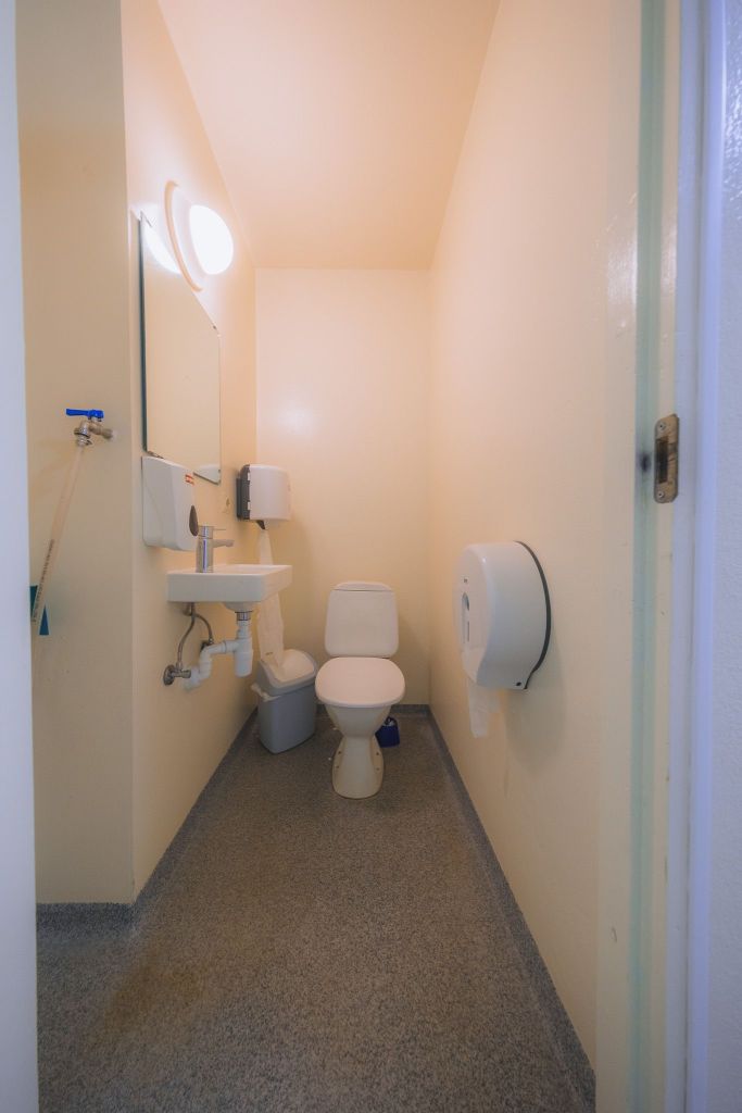 Small, clean restroom with beige walls and gray flooring. Features a toilet, sink with mirror, hand dryer, and toilet paper dispenser.