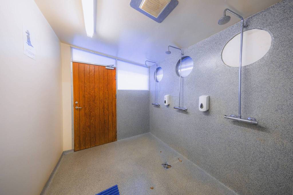 Simple shower room with three showerheads on a gray wall, round windows above, wood door at the end, and dispensers for soap.