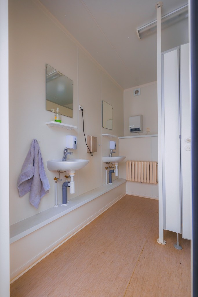 Clean and minimalistic restroom with two white sinks, mirrors, and soap dispensers on a light wood floor. A towel hangs beside one sink.