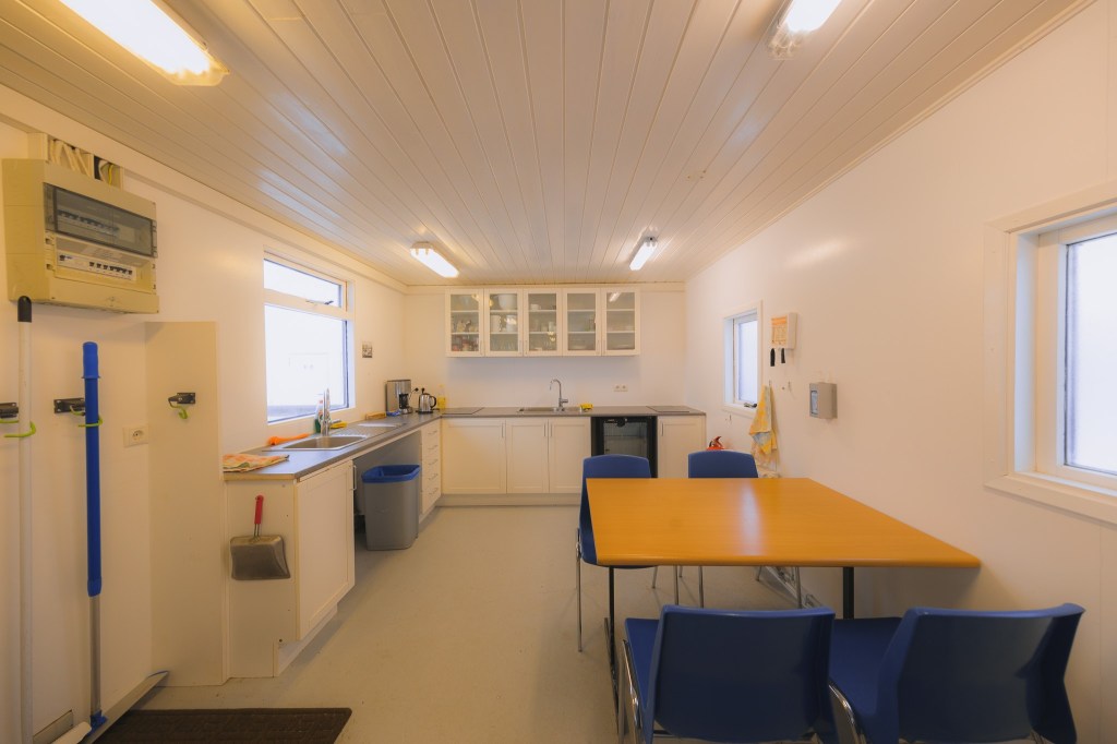 Bright, tidy kitchen with white walls and ceiling. Contains a sink, countertop, cabinets, blue chairs around a wooden table, and large windows providing ample light.
