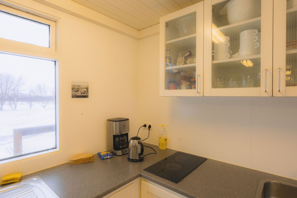 Bright kitchen corner with a window view of snowy trees. Coffee maker and kettle on the countertop. White cabinets hold mugs and bowls.