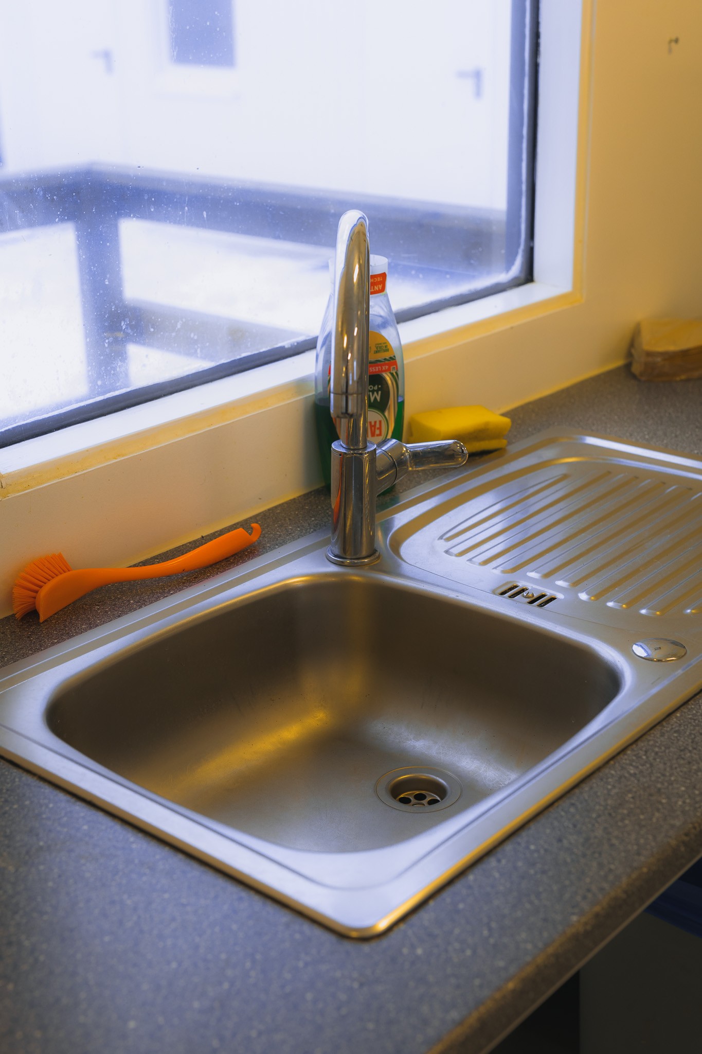 Stainless steel kitchen sink with a faucet, sponge, and dish soap near a window. A bright orange scrub brush is on the counter.