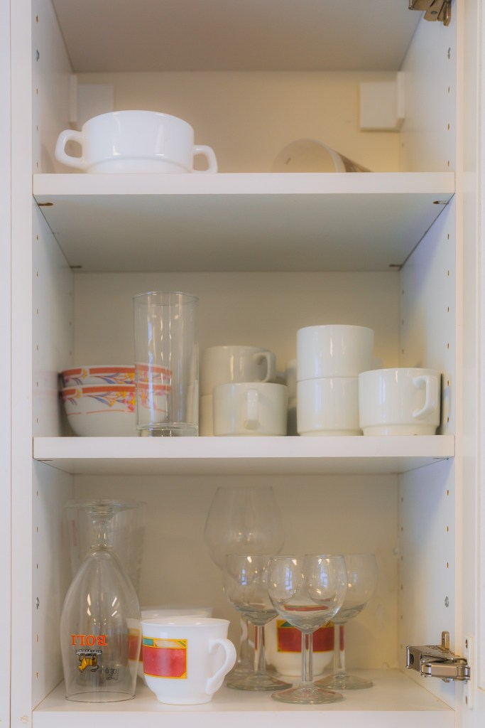 Open kitchen cupboard with neatly arranged dishes. Top shelf: white bowls and cups. Middle: stacked cups and glasses. Bottom: glasses and mug.